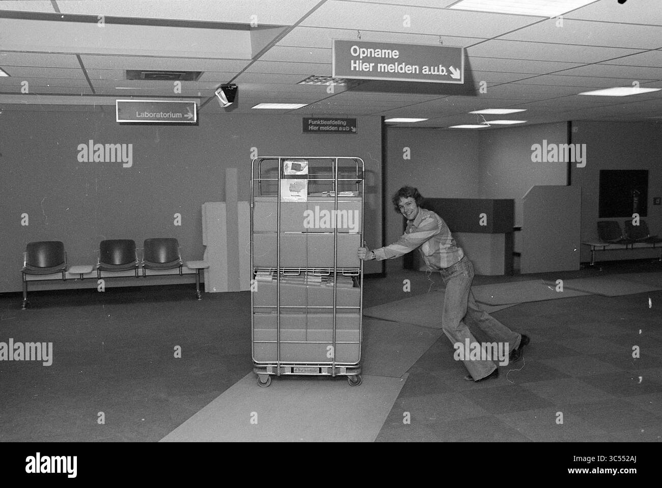 Déménagement. Joseph ZH, Relocation, 06-02-1978 Whizgle News, Dutch Desk, pays-Bas, 1950-2000 Une personne pousse un chariot chargé de boîtes dans une aire d'attente vide, entourée de chaises et de panneaux indiquant les différentes sections d'une installation. Banque D'Images