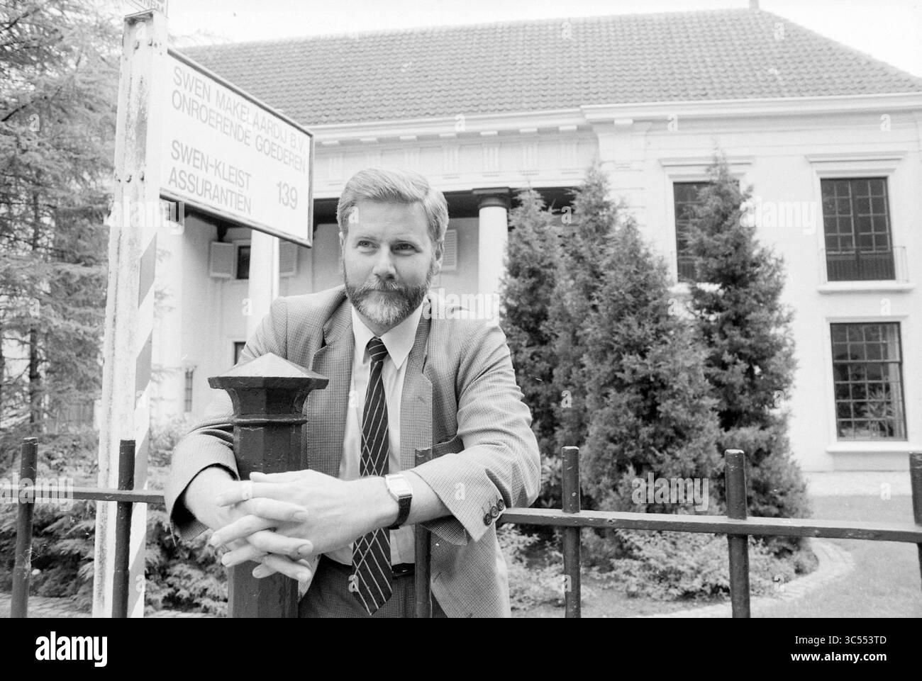 Agent immobilier en face de son bureau Whizgle News, Dutch Desk, pays-Bas, 1950-2000 Un homme se tient sans heurt près d'une clôture, penché les bras croisés. Il porte une barbe soignée et porte un costume avec une cravate. Derrière lui, un bâtiment présente une architecture classique, avec de hautes colonnes et un panneau indiquant son nom et son adresse. Une végétation luxuriante entoure la scène, ajoutant à l'atmosphère sereine. Banque D'Images