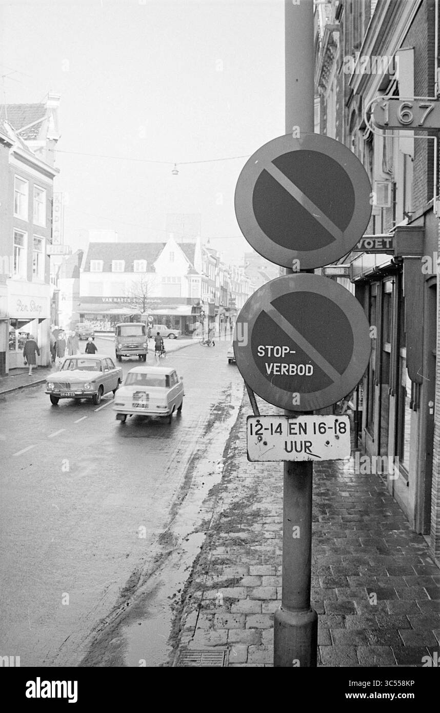 Interdiction de stationnement et d'arrêt, Haarlem, Grote Houtstraat, pays-Bas, 00-00-1965 Whizgle News, Dutch Desk, pays-Bas, 1950-2000 Une scène de rue présente un panneau indiquant l'interdiction de stationnement affiché en évidence sur un poteau, avec deux icônes circulaires indiquant les restrictions. La route est bordée de bâtiments, et quelques voitures anciennes peuvent être vues conduire au loin, ajoutant une atmosphère nostalgique au cadre urbain. Banque D'Images