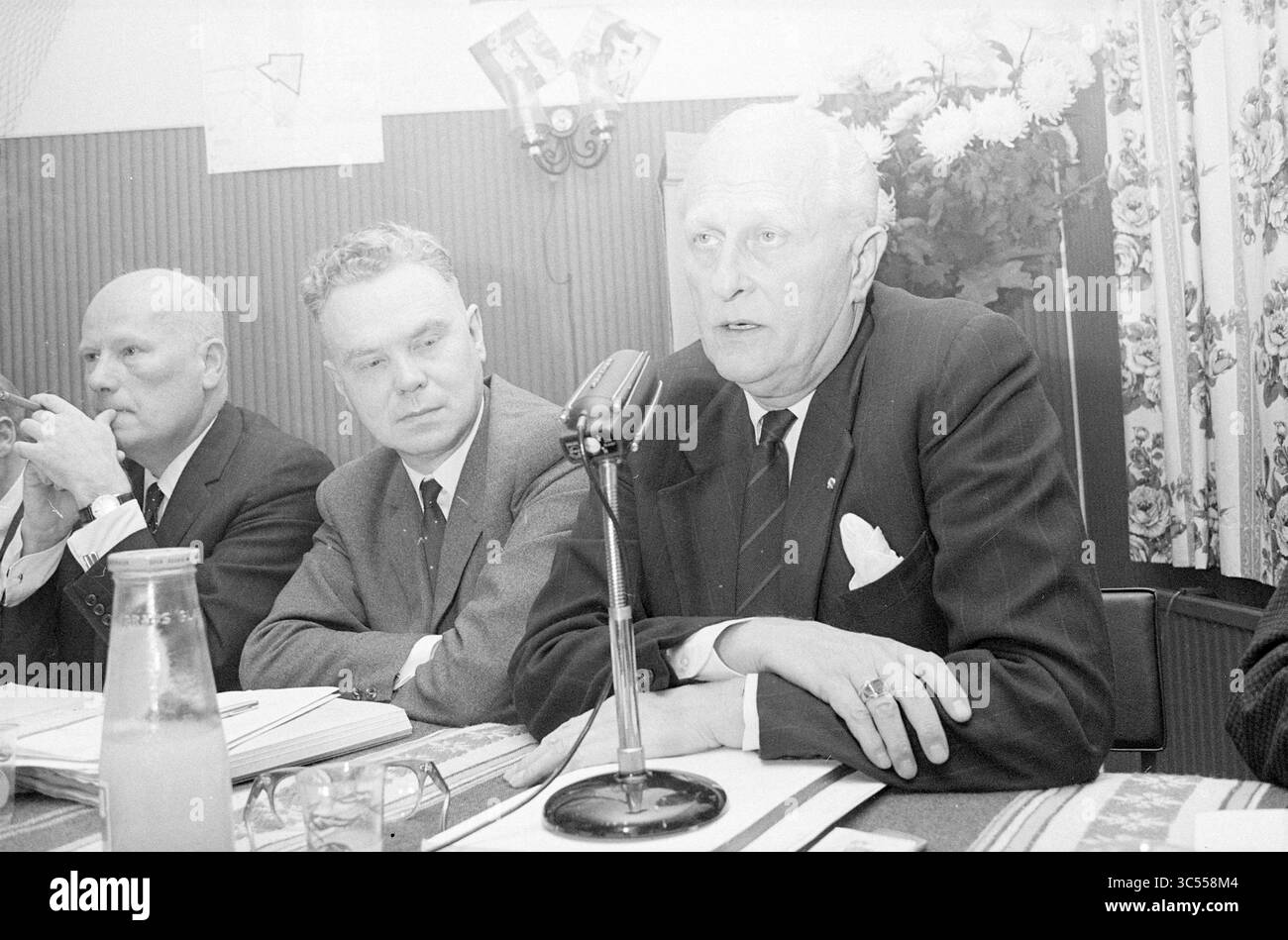 Teach-In Noise pollution Schiphol, Schiphol, 02-11-1967 Whizgle News, Dutch Desk, pays-Bas, 1950-2000 Un groupe de trois hommes est assis à une table pendant une réunion, un homme parlant dans un microphone. Les individus semblent engagés dans la discussion, et il y a des documents et une bouteille devant eux, avec une toile de fond de rideaux à motifs floraux. Banque D'Images