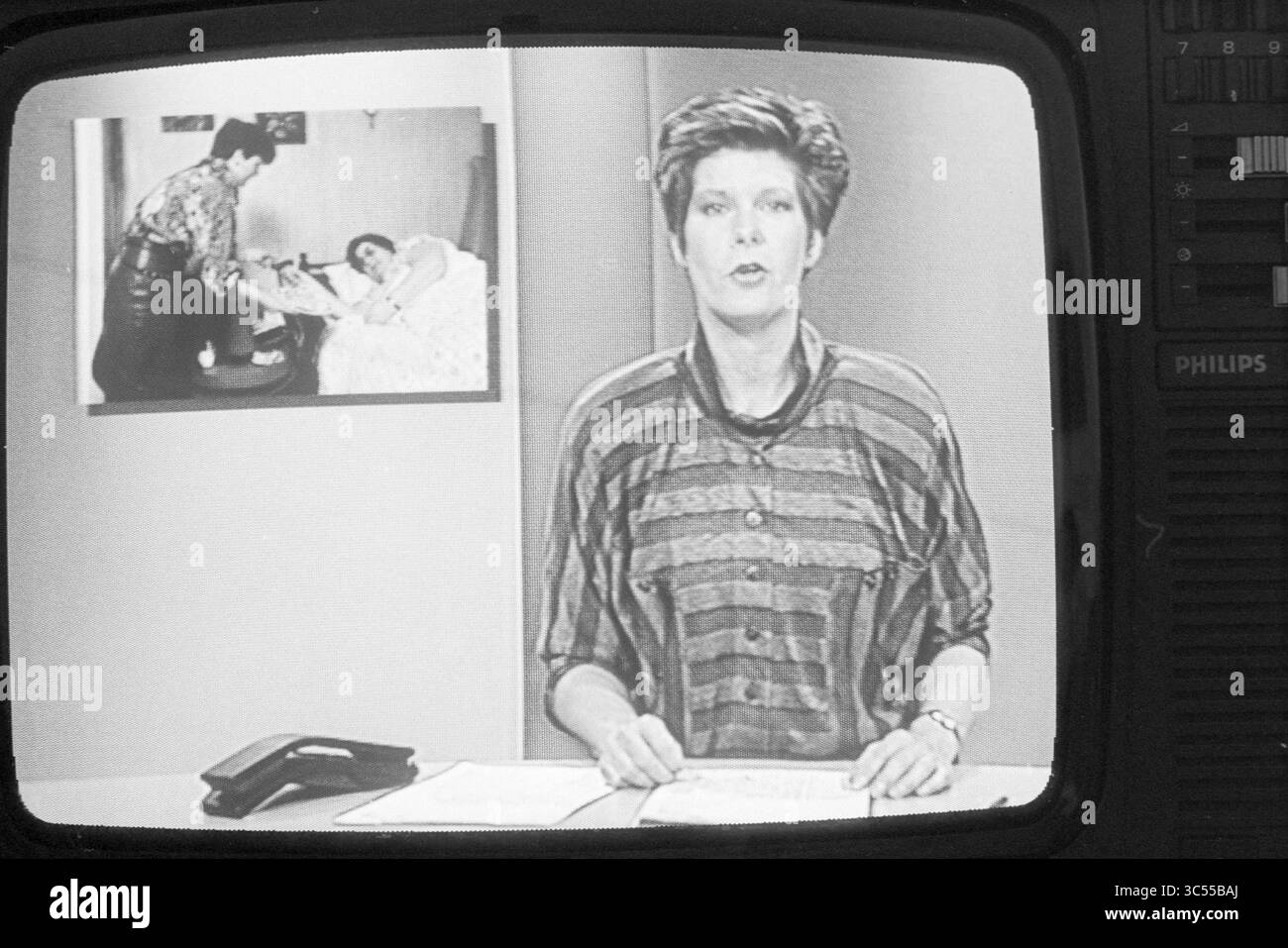 Maartje van Weegen nos news, télévision, 26-07-1988 Whizgle News, Dutch Desk, pays-Bas, 1950-2000 Un présentateur présente un reportage dans un studio, avec une image encadrée de deux individus dans un cadre domestique visible en arrière-plan. L'ancre est habillée d'un dessus rayé et est positionnée à un bureau avec des papiers et un téléphone. Banque D'Images