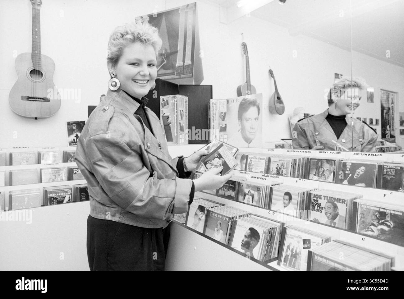CD shop, Kennemerlaan, IJmuiden, boutiques et kiosques, IJmuiden, Kennemerlaan, pays-Bas, 06-11-1987 Whizgle News, Dutch Desk, pays-Bas, 1950-2000 Une femme avec une coupe de cheveux élégante parcourt une collection d'albums de musique, tenant un CD tout en souriant à la caméra. Derrière elle, divers disques et une guitare sont affichés sur le mur, ajoutant une ambiance vintage à la scène. Banque D'Images