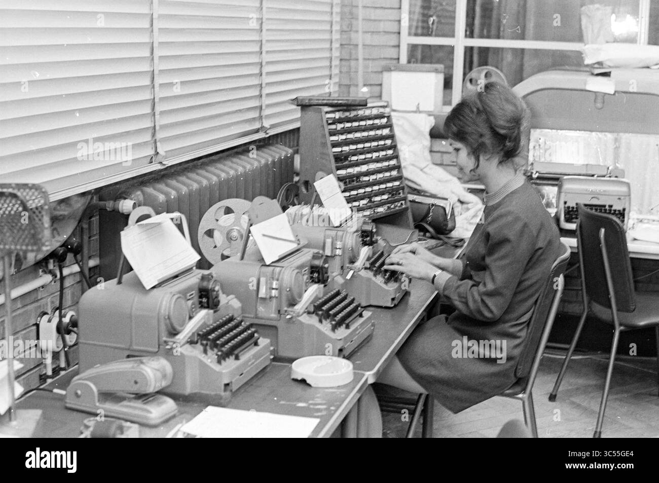 Girl Behind telex machine, Persons, 16-12-1969 Whizgle News, Dutch Desk, pays-Bas, 1950-2000 Une femme vêtue d'une tenue intelligente s'assoit à un bureau, tapant sur une machine à écrire vintage entourée de plusieurs autres machines similaires. Les papiers et les notes sont éparpillés, et le décor présente de grandes fenêtres avec stores et un radiateur, créant une atmosphère de bureau animée. Banque D'Images Girl Behind telex machine, Persons, 16-12-1969 Whizgle News, Dutch Desk, pays-Bas, 1950-2000 Une femme vêtue d'une tenue intelligente s'assoit à un bureau, tapant sur une machine à écrire vintage entourée de plusieurs autres machines similaires. Les papiers et les notes sont éparpillés, et le décor présente de grandes fenêtres avec stores et un radiateur, créant une atmosphère de bureau animée. Banque D'Images