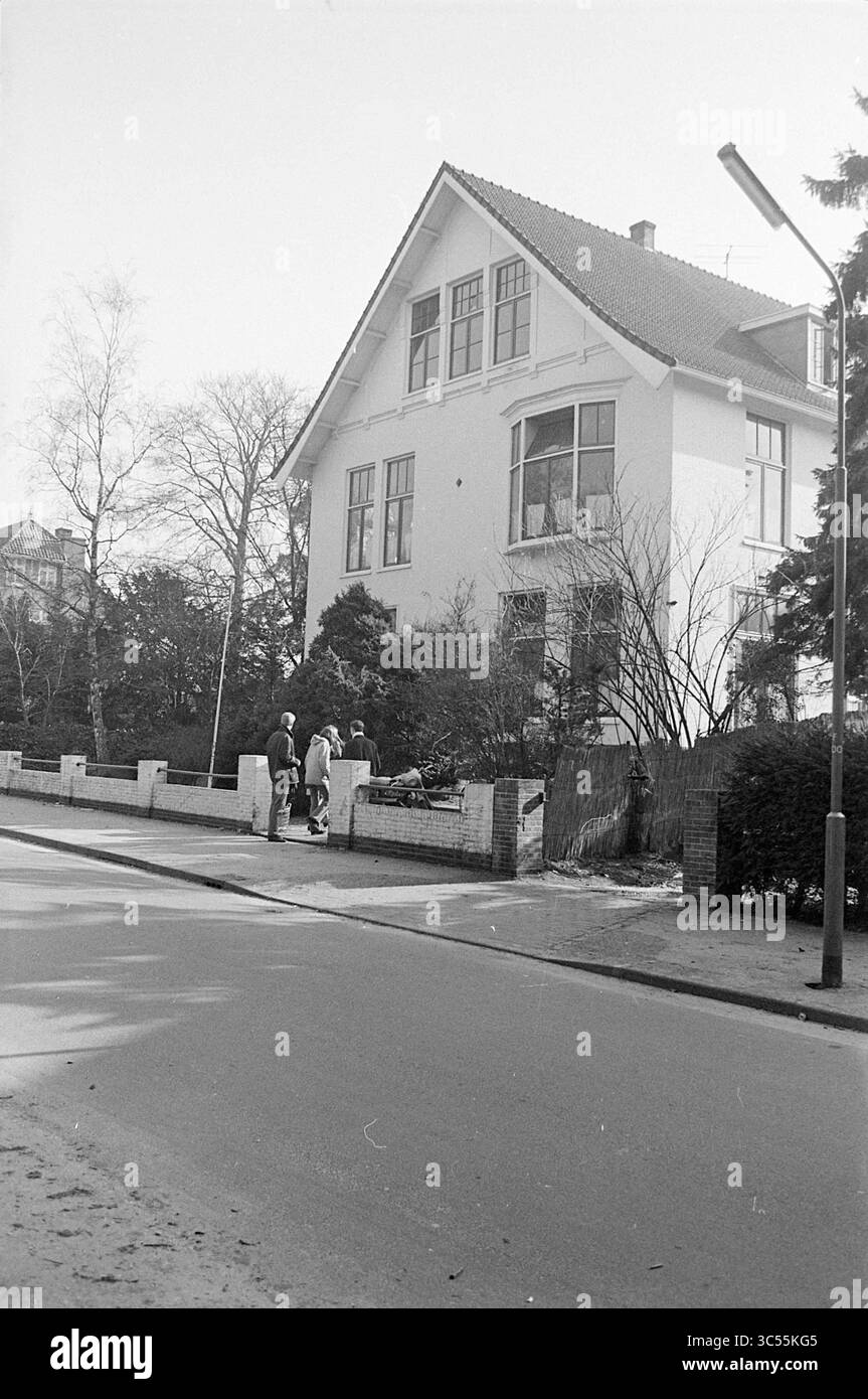 Villa extérieure, 00-03-1969 Whizgle News, Dutch Desk, pays-Bas, 1950-2000 Une maison de trois étages avec un toit à pignon distinctif se dresse bien en évidence le long d'une rue calme, entourée d'arbres et de buissons. Un petit groupe de personnes discute à l'extérieur de la clôture, tandis qu'un lampadaire se tient à proximité, projetant une ombre sur la route. Banque D'Images