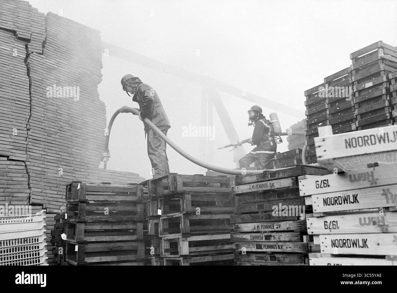Hangar à ampoules à feu fa. Willemsen Hillegom, pompiers, Hillegom, 02-03-1980 Whizgle News, Dutch Desk, pays-Bas, 1950-2000 les pompiers en équipement de protection travaillent à éteindre un incendie au milieu de piles de palettes en bois, avec de la fumée qui circule autour d'eux alors qu'ils pulvérisent de l'eau à partir d'un tuyau. Banque D'Images