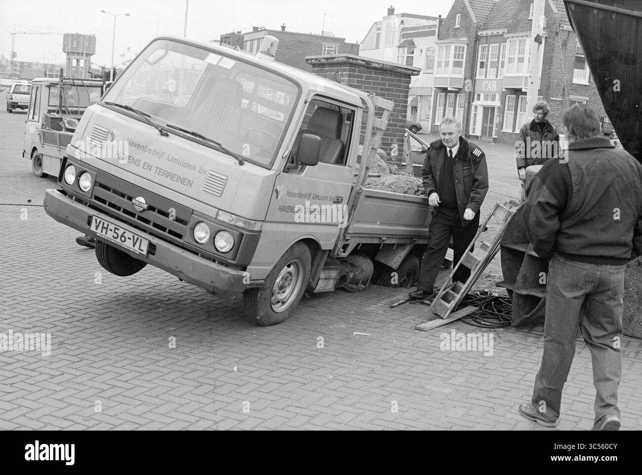 Car in hole, IJm., IJmuiden, pays-Bas, 31-01-1994 Whizgle News, Dutch Desk, pays-Bas, 1950-2000 Un petit camion de livraison est partiellement incliné dans un chantier de construction, un côté reposant sur le sol. Deux travailleurs sont à proximité, observant la situation, tandis que les environs présentent un mélange de bâtiments et d'équipements. Banque D'Images