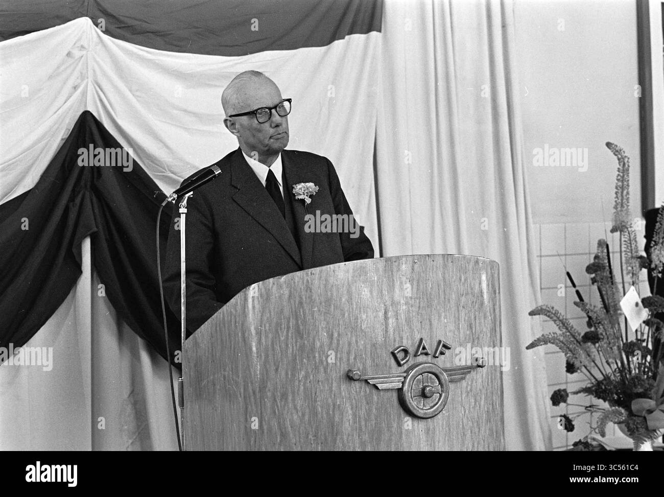 Discours, N.A.M nouvelle société automobile Whizgle News, Dutch Desk, pays-Bas, 1950-2000 Un homme en costume se tient sur un podium orné d'un logo, prononçant un discours. Il porte des lunettes et une boutonnière sur son revers, avec un fond de tissu drapé en rouge et blanc, suggérant un événement formel ou une cérémonie. Banque D'Images