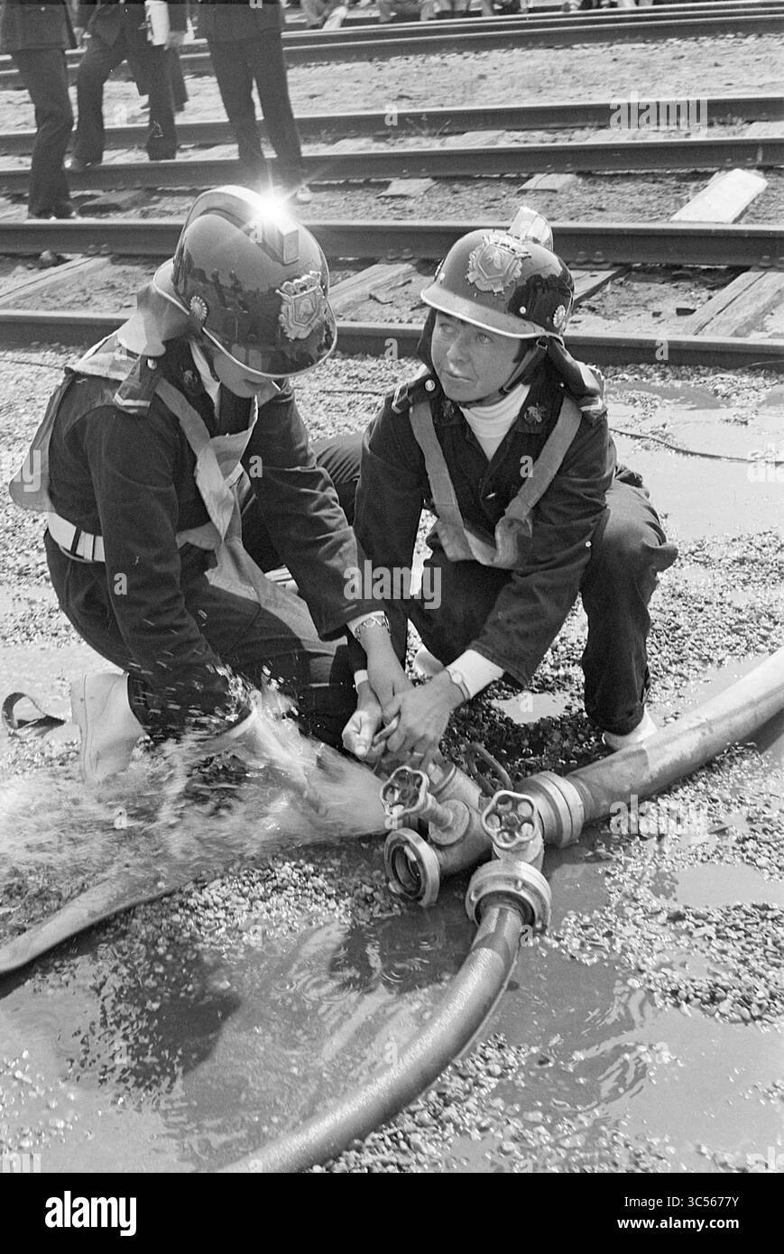 Pompiers compétitions équipe féminine Haarlem, incendies, pompiers, compétitions, 15-05-1976 Whizgle News, Dutch Desk, pays-Bas, 1950-2000 deux pompiers travaillent ensemble pour connecter un gros tuyau d'incendie, axé sur leur tâche de pulvérisation d'eau par la buse. Ils portent des casques et des équipements de protection, illustrant le travail d'équipe et la détermination dans un effort de lutte contre l'incendie. La toile de fond présente des voies ferrées, suggérant un cadre industriel. Banque D'Images