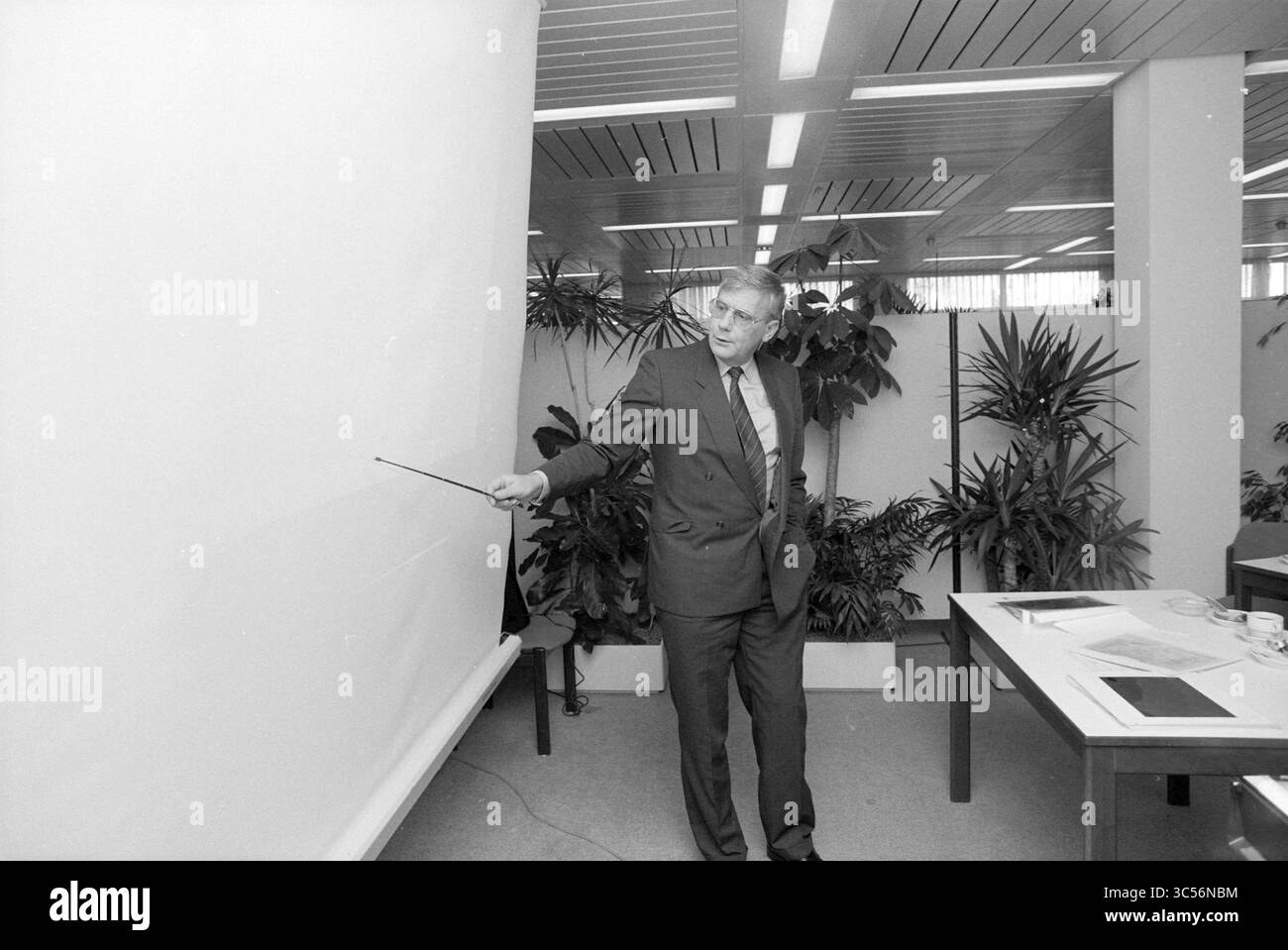 Staff Members course, N.A.M., 23-10-1986 Whizgle News, Dutch Desk, pays-Bas, 1950-2000 Un homme en costume se tient confiant, tenant un pointeur alors qu'il fait un geste vers un écran de présentation vide. Autour de lui se trouvent des plantes en pot, et une table est dressée avec des documents et du matériel, suggérant un cadre professionnel propice à une réunion ou une conférence. Banque D'Images