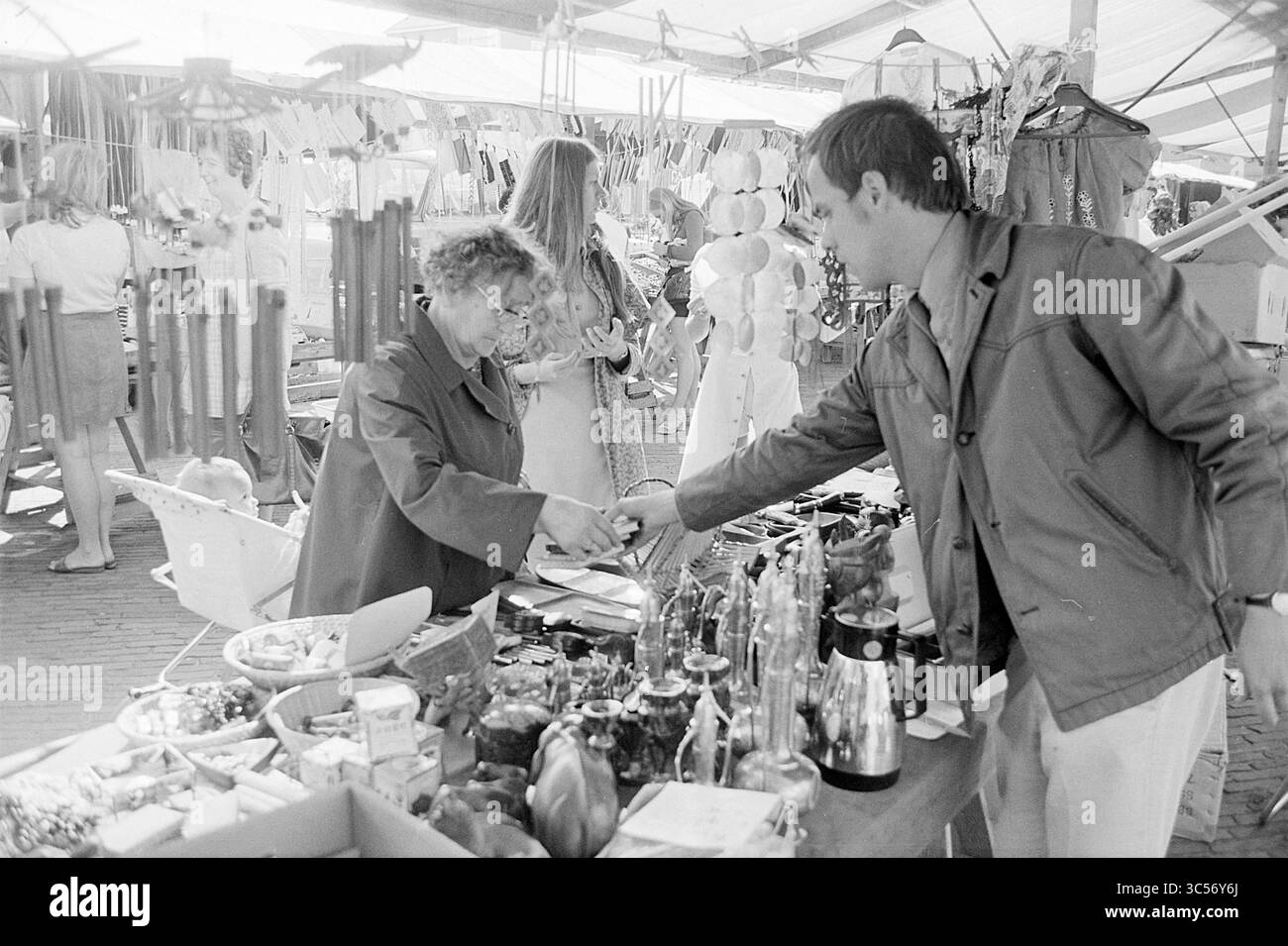 Rapport marché, IJmuiden, rapport, IJmuiden, Markt, pays-Bas, 31-08-1972 Whizgle News, Dutch Desk, pays-Bas, 1950-2000 Une scène de marché animée capture un moment d'interaction entre un vendeur et un client, entouré d'un éventail de produits colorés et d'artisanat artisanal, avec d'autres acheteurs qui naviguent en arrière-plan. Banque D'Images