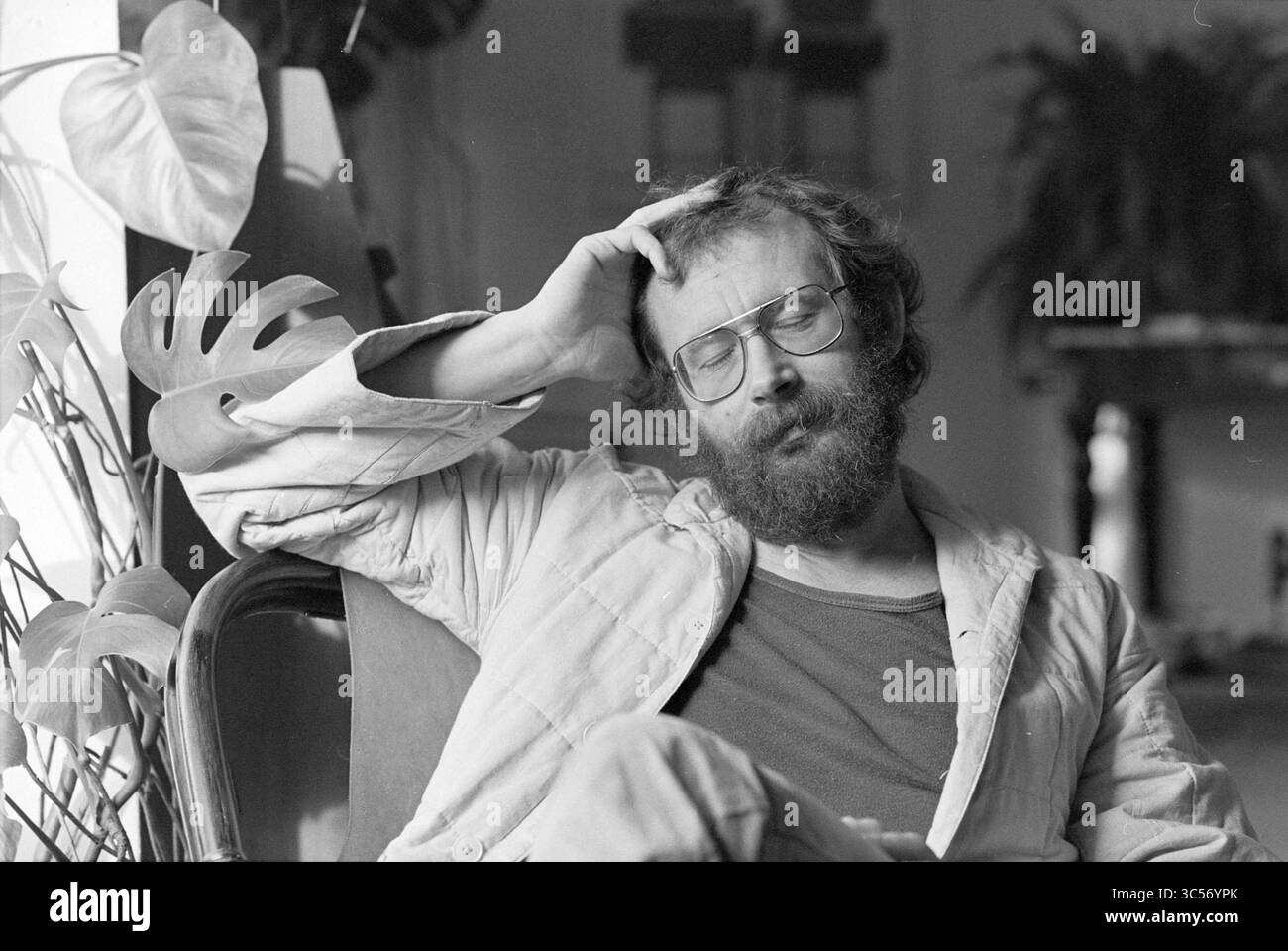 Portrait de Hans Dorrestein, Herwijnen (parolier), personnes écrivant, 01-11-1978 Whizgle News, Dutch Desk, pays-Bas, 1950-2000 Un homme contemplatif avec des lunettes et une barbe est assis détendu, reposant sa main sur sa tête tout en étant perdu dans ses pensées. Une tenue décontractée et un éventail de verdure en arrière-plan créent une atmosphère sereine. Banque D'Images