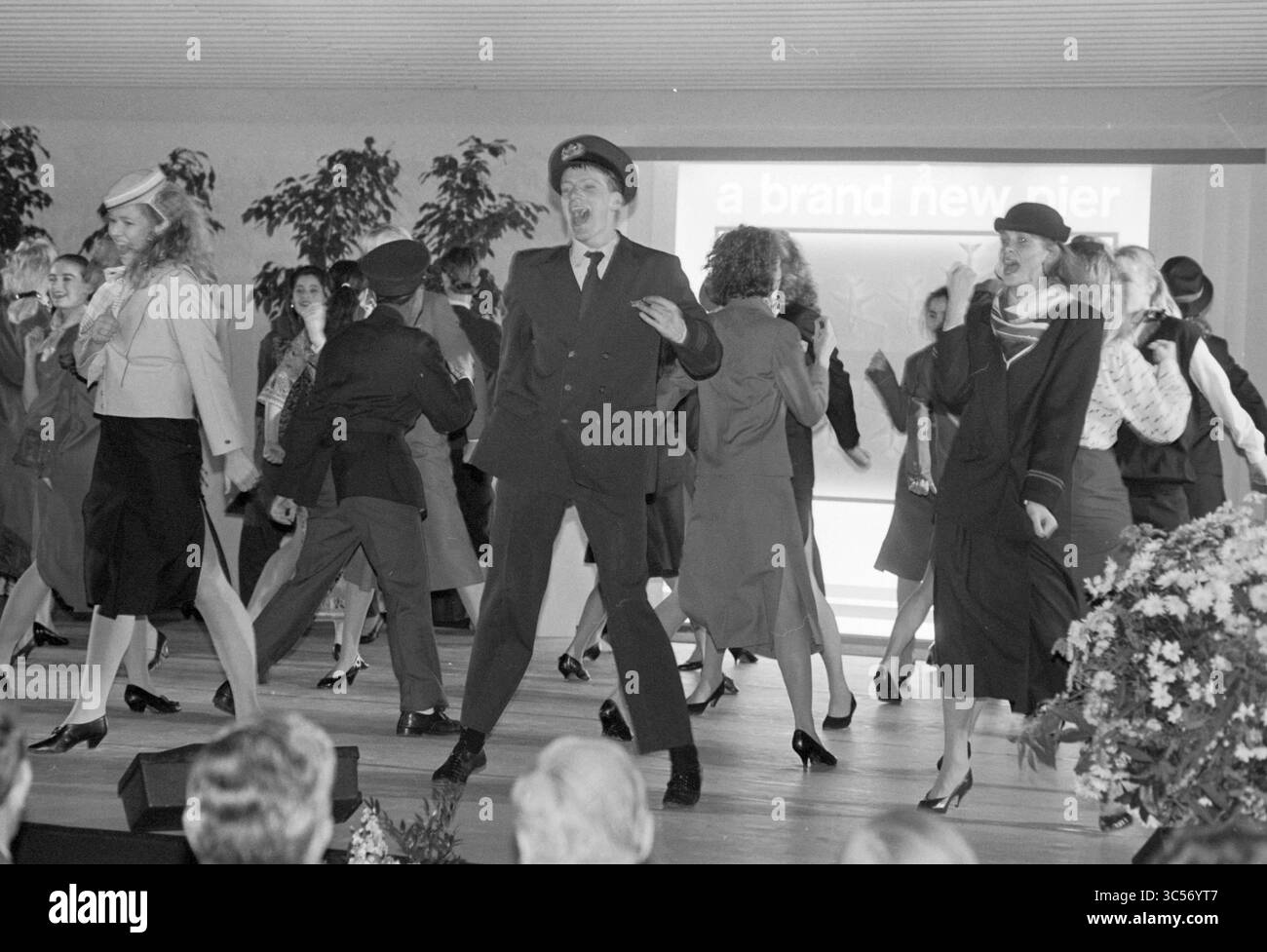 Ouverture de C-Pier Schiphol, Schiphol, 13-12-1987 Whizgle News, Dutch Desk, pays-Bas, 1950-2000 Une performance animée mettant en vedette un groupe de danseurs habillés dans des vêtements élégants, mettant en vedette des mouvements synchronisés et des sourires. La toile de fond comprend un grand écran avec le texte « a flambant neuf jetée », suggérant un thème de célébration. La scène est remplie d’énergie et d’enthousiasme, avec un accent sur la mode et la chorégraphie. Banque D'Images