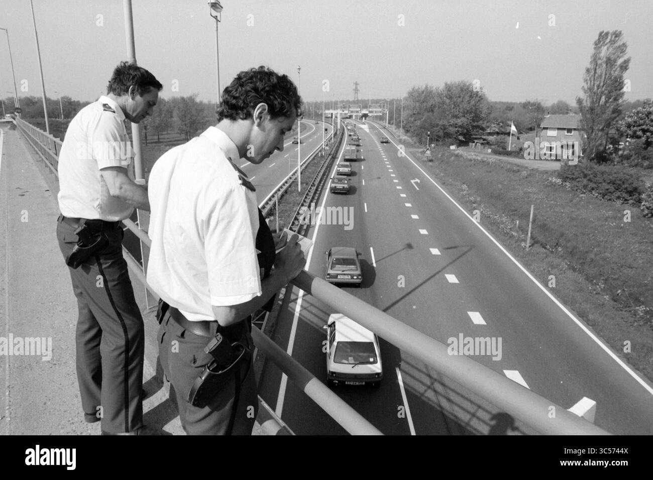 Contrôle de la police à l'embouteillage Velsertunnel, police, Velsertunnel, Beverwijk, Rijksweg A22, pays-Bas, 09-05-1989 Whizgle News, Dutch Desk, pays-Bas, 1950-2000 deux policiers se tiennent sur un pont, observant la circulation en contrebas. Une ligne de voitures se déplace le long d'une autoroute très fréquentée, flanquée de verdure et de bâtiments voisins. Les agents semblent concentrés, enregistrant ou surveillant peut-être le flux des véhicules. Banque D'Images