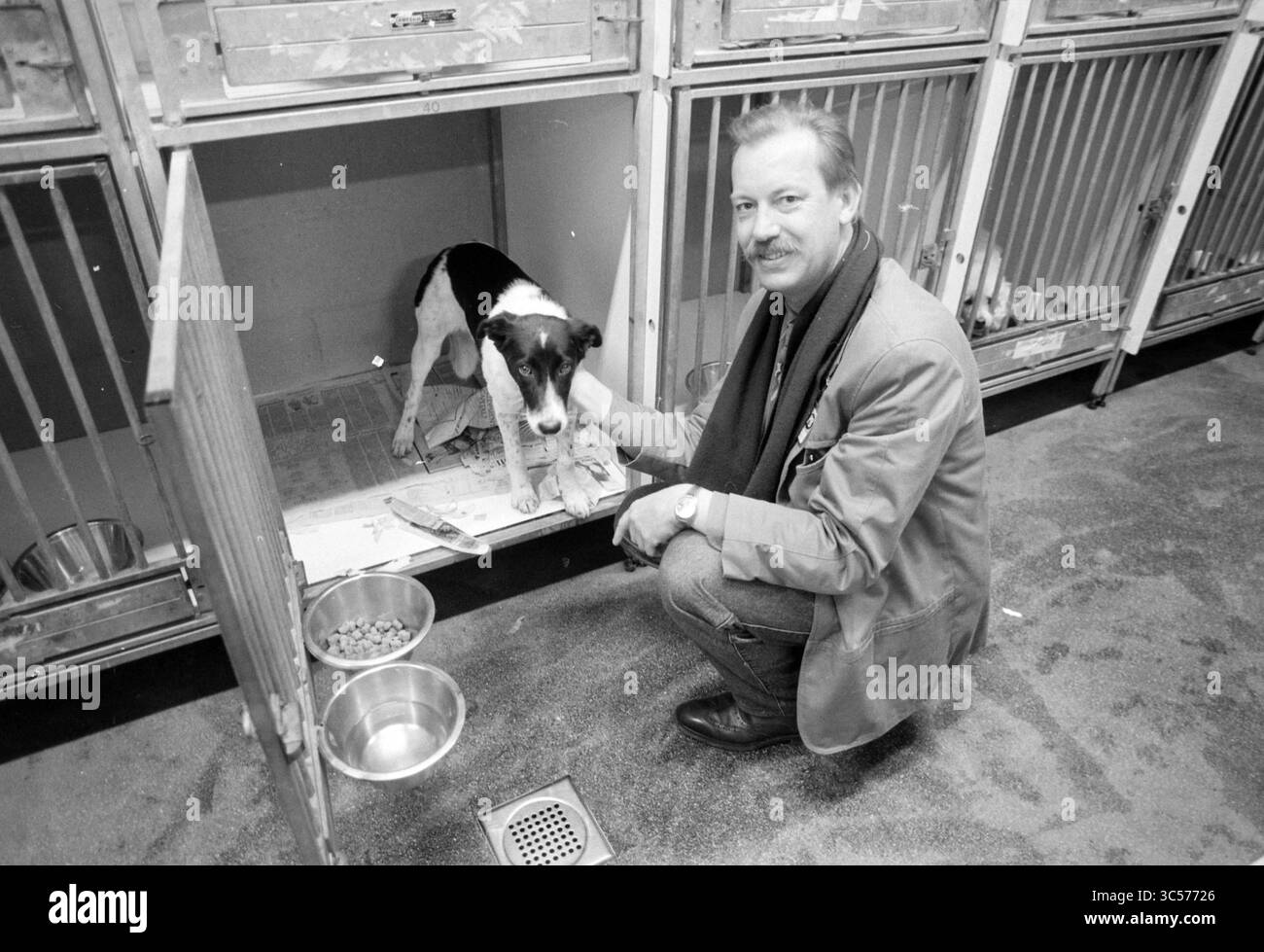 Refuge pour animaux, 00-04-1991 Whizgle News, Dutch Desk, pays-Bas, 1950-2000 Un homme s'agenouille à côté d'un chien dans un chenil, offrant confort et compagnie. Le chien semble attentif et curieux, tandis que le chenil dispose de bols de nourriture et d'eau à proximité. Banque D'Images