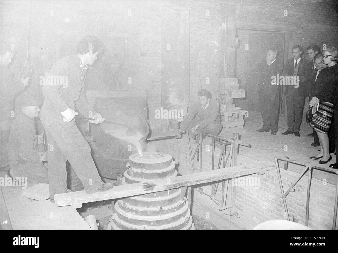 B et W visiter la fonderie de cloches à Asten, horloges et montres, IJmuiden, Velsen, 15-10-1964 Whizgle News, Dutch Desk, pays-Bas, 1950-2000 Un groupe de personnes observe en tant que travailleur façonne habilement des matériaux fondus dans un cadre d'atelier historique, mettant en valeur l'artisanat traditionnel et l'art du travail des métaux. Banque D'Images