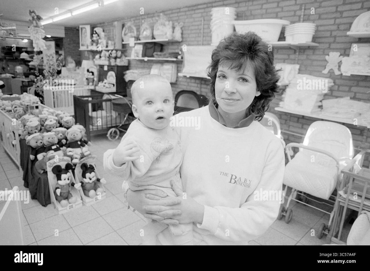 Marian Goedhart de Baby bussines, magasins et kiosques, 27-01-1989 Whizgle News, Dutch Desk, pays-Bas, 1950-2000 Une femme tient un bébé debout dans un magasin rempli d'articles pour bébés et de jouets en peluche, mettant en valeur une atmosphère chaleureuse de soins pour enfants et d'articles parentaux essentiels. Banque D'Images