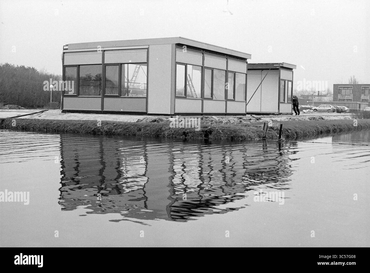 Construction d'un bâtiment d'urgence localement à côté du canal, 00-03-1992 Whizgle News, Dutch Desk, pays-Bas, 1950-2000 A Modern, structure minimaliste se trouve sur une petite île, entourée d'eau, reflétant ses lignes nettes et son design dans la surface calme. En arrière-plan, des éléments industriels font allusion à un environnement urbain proche. Banque D'Images