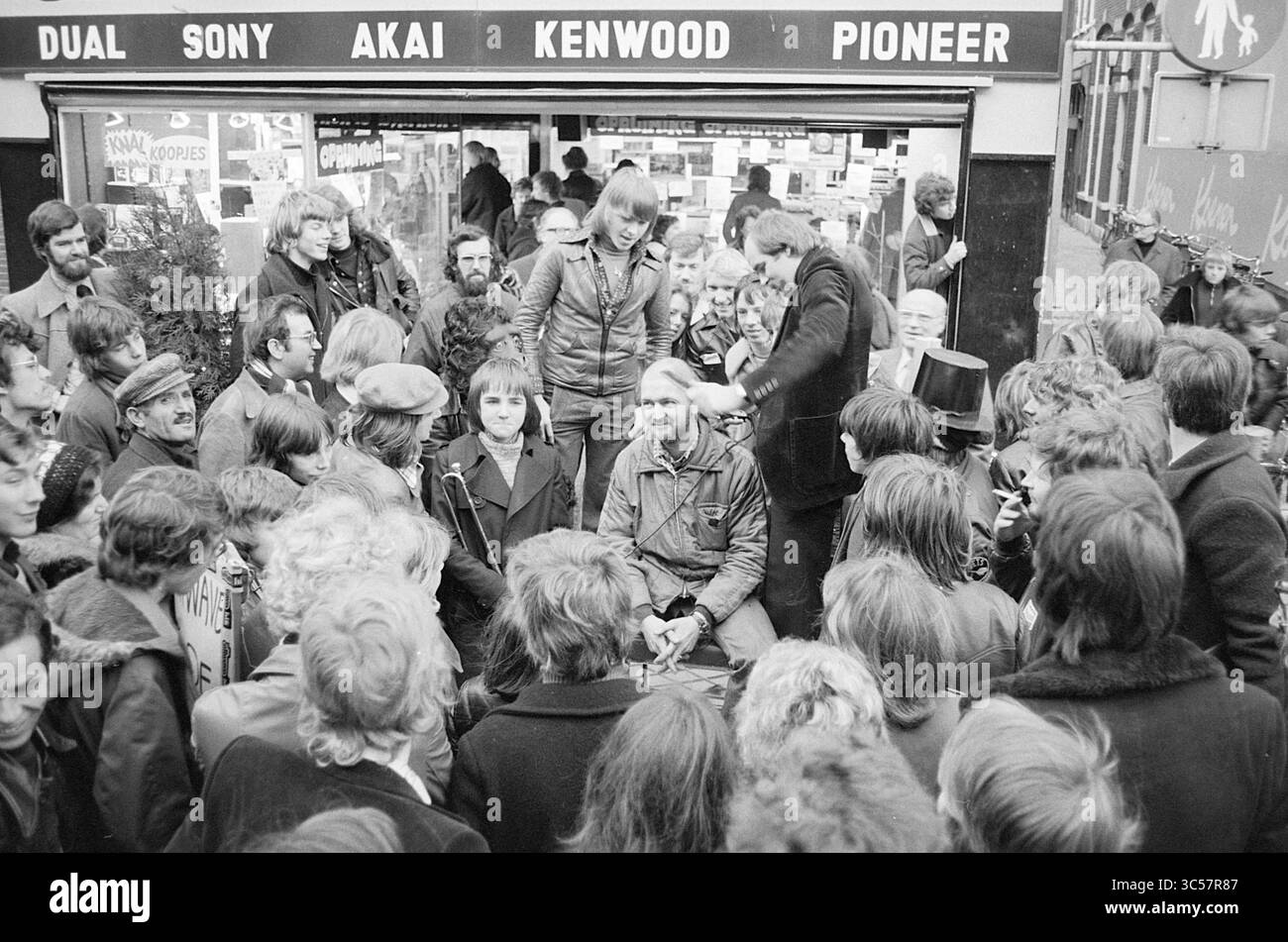 Sale All Wave Haarlem., Sale, Haarlem, Grote Houtstraat, pays-Bas, 15-01-1976 Whizgle News, Dutch Desk, pays-Bas, 1950-2000 Une foule animée entoure un groupe d'individus assis devant un magasin d'électronique, mettant en avant un moment d'engagement et d'excitation. Des personnes de différents âges sont rassemblées, beaucoup montrant des expressions de curiosité et d'enthousiasme, alors qu'elles regardent une démonstration ou un événement en cours. La vitrine présente des marques de renom comme Sony, Akai et Pioneer, ce qui laisse entendre un accent particulier sur l'équipement audio et la technologie. Banque D'Images