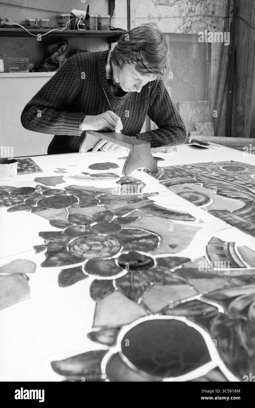 GlazierJan Nelis, vitraux, soufflage de verre, verre en général, 19-11-1980 Whizgle News, Dutch Desk, pays-Bas, 1950-2000 un artiste peint méticuleusement des motifs floraux complexes sur une grande toile, concentrés et immergés dans leur métier. L'espace de travail environnant laisse entrevoir un environnement créatif, rempli d'outils et de matériaux pour l'expression artistique. Banque D'Images