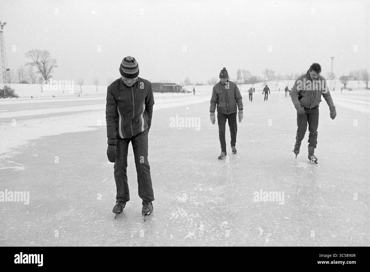 Leçons de patinage à Nova Zembla, Spaarndam, patinage, Spaarndam, 15-01-1985 Whizgle News, Dutch Desk, pays-Bas, 1950-2000 Un groupe de patineurs glisse sur une surface glacée, vêtus chaudement pour les conditions froides. Une personne se penche en avant, en se concentrant sur ses foulées, tandis que d'autres patinent à distance, entourés d'un paysage hivernal d'arbres et de structures faibles en arrière-plan. Banque D'Images