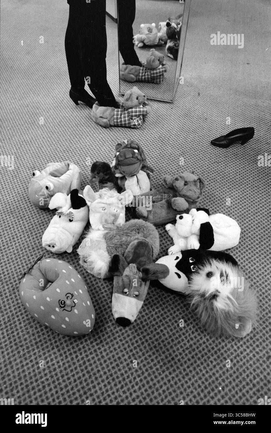 Pantoufles, engouement avec les animaux, 01-12-1991 Whizgle News, Dutch Desk, pays-Bas, 1950-2000 Une collection de chaussons et de jouets en peluche colorés est dispersée sur un tapis à motifs, avec un reflet des jambes d'une personne visible dans un miroir voisin. Une chaussure noire à talons hauts contraste avec l'assortiment fantaisiste. Banque D'Images