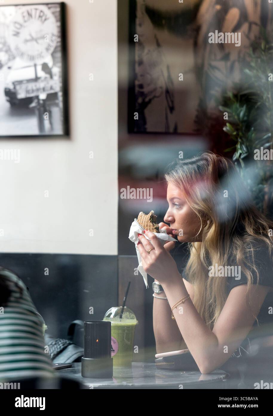 Jeune femme mangeant dans un café londonien, vue à travers la fenêtre, avec des reflets et un flou : style de vie urbain et photographie de rue franche. Banque D'Images