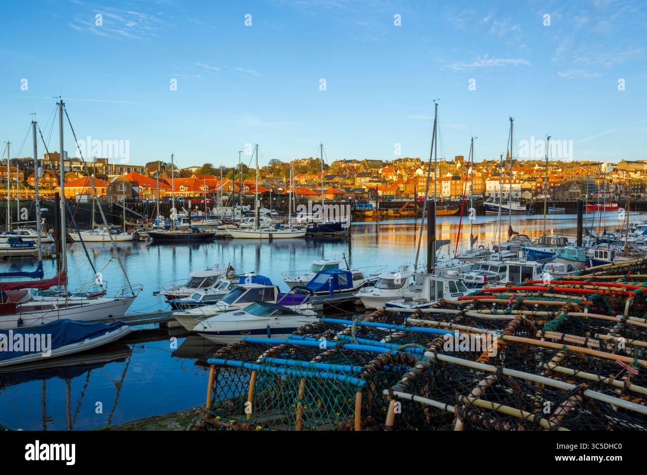 Vue sur la lumière dorée du matin à travers la marina de Whitby avec des bateaux amarrés et des casiers à homard au premier plan et des bâtiments à flanc de colline en arrière-plan Banque D'Images