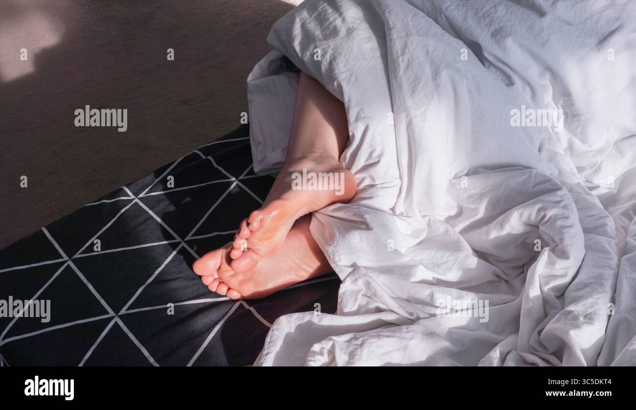 La lumière du soleil illuminant les pieds de la femme sortant de la couverture blanche froissée sur le lit avec un motif géométrique, transmettant des concepts de repos, de relaxation et de c. Banque D'Images