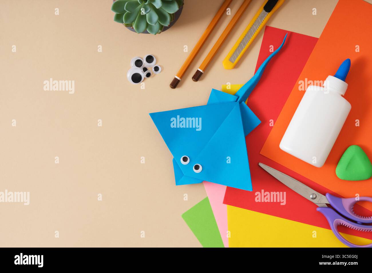 Table de travail pour papier artisanal feuilles colorées, colle, ciseaux, crayons, couteau de papeterie. Jouet origami finit Stingray. Fond beige, espace pour votre Banque D'Images