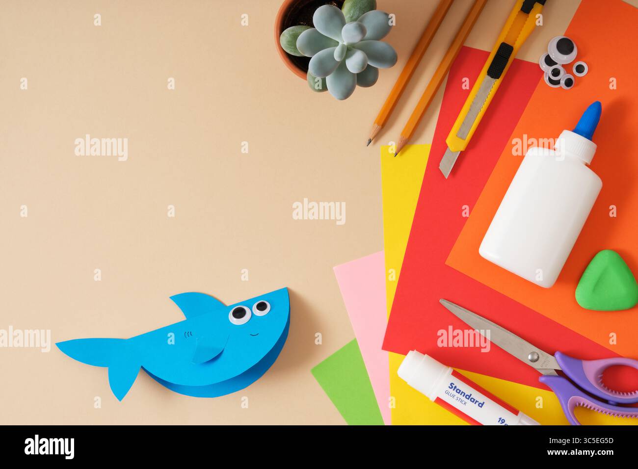 Requin jouet en papier fini. Fond beige, espace pour le texte. Table de travail pour papier artisanal feuilles colorées, colle, ciseaux, crayons, couteau de papeterie. Banque D'Images