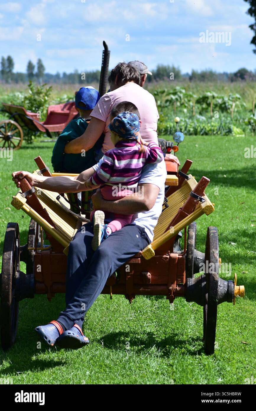 Famille avec deux enfants à cheval sur un chariot en bois jaune tiré par un tracteur à travers la campagne verdoyante. Une joyeuse expérience de liaison en plein air sous le bleu Banque D'Images