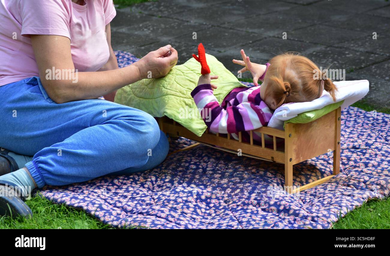 Grand-mère et petite fille assis à l'extérieur sur une couverture à motifs, jouant avec un lit de jouet et des articles faits à la main. Moment de liaison chaud dans la cour arrière ensoleillée s Banque D'Images