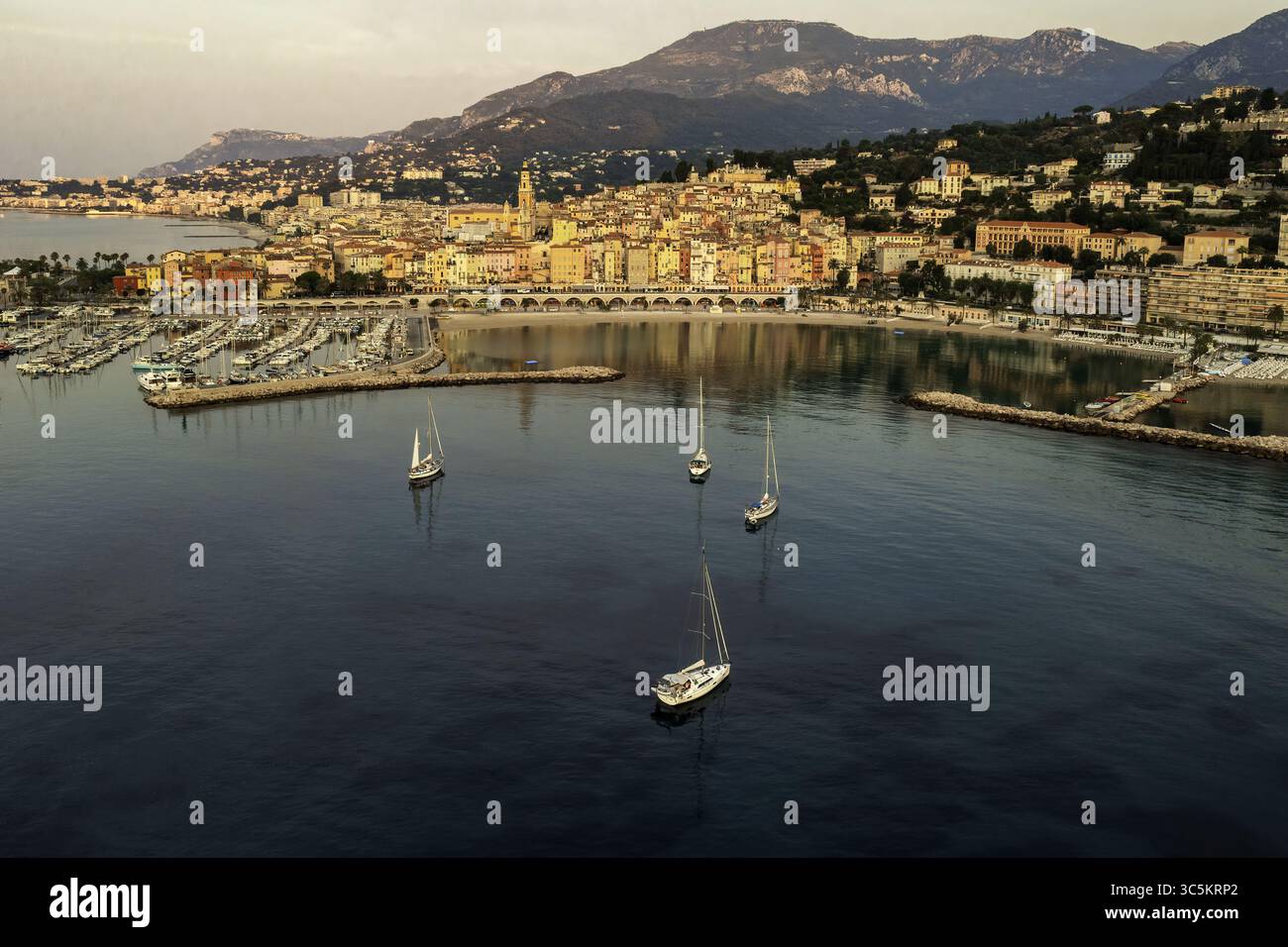 Vue aérienne du port rempli de mâts de voiliers réfléchissant la lumière, niché contre les bâtiments vibrants et les montagnes lointaines, Menton, Provence-Alpes-Côte d'Azur, France. Banque D'Images
