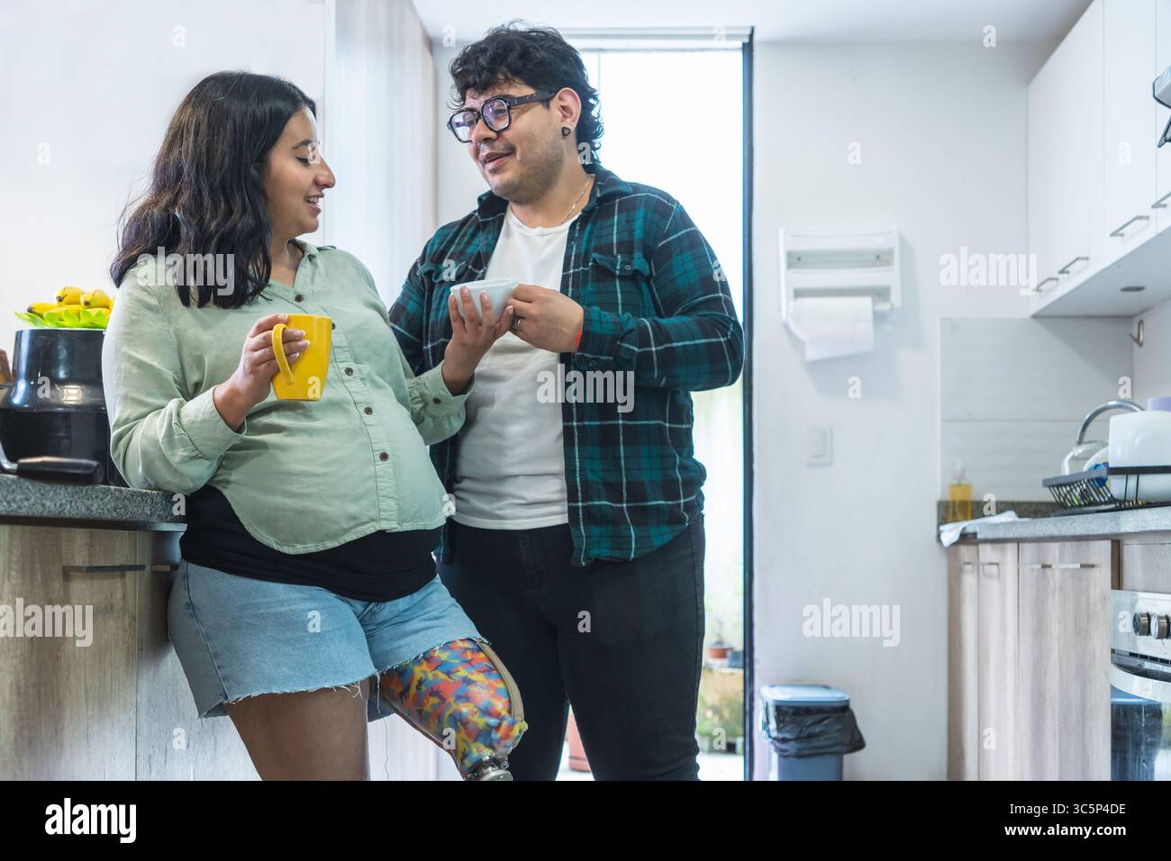 Couple heureux, femme enceinte avec jambe prothétique et mari, partageant un moment ensemble tout en buvant du café dans leur cuisine Banque D'Images