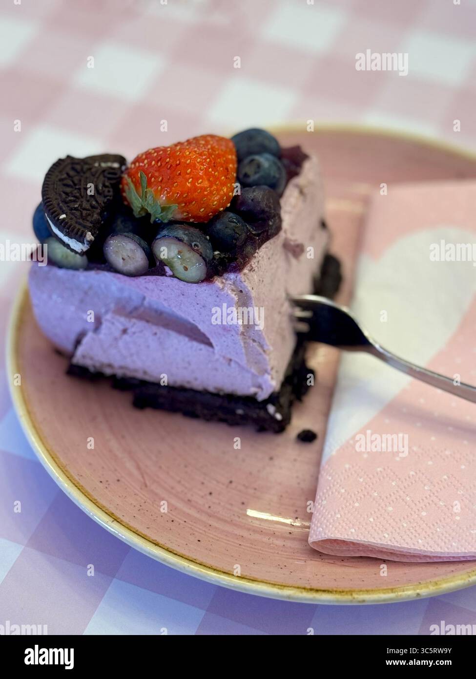 Une tranche de gâteau mousse aux baies avec une base de biscuit au chocolat, garnie de bleuets, une fraise et un Oreo, servi sur une assiette rose avec un coeur na Banque D'Images