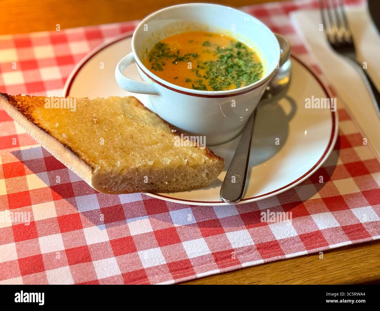 Un bol de soupe de tomates garni de persil servi avec une tranche de pain à l'ail sur une assiette blanche posée sur une nappe à carreaux rouge à Lübeck, en Allemagne. - Image de stock capturée avec un smartphone