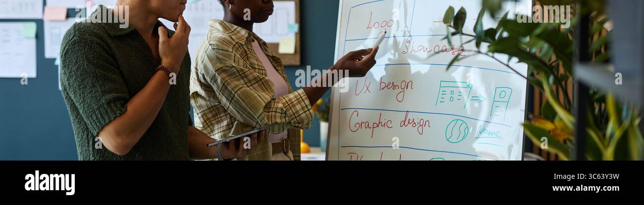Jeune homme asiatique adulte tenant la tablette, debout à côté de la jeune femme noire adulte pointant vers le tableau blanc discutant des concepts de design UX dans l'espace de travail de bureau moderne pendant la réunion créative Banque D'Images