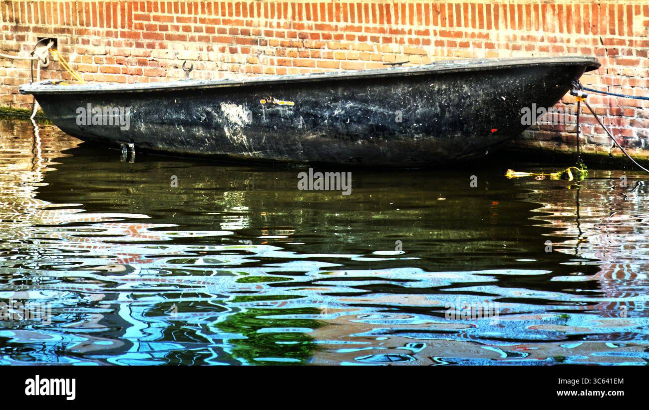 Petit canot noir amarré à côté d'un mur de briques altérées, se balançant doucement dans les eaux calmes du paisible canal, créant des ondulations douces Banque D'Images