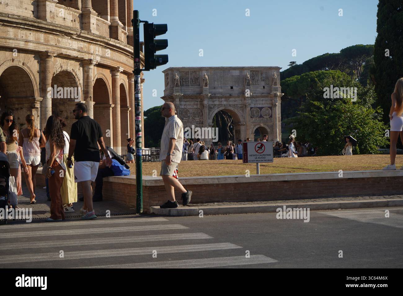 Les touristes marchent près du Colisée et de l'Arc de Constantin sous un ciel dégagé. Un feu de circulation et un passage pour piétons sont visibles. Banque D'Images