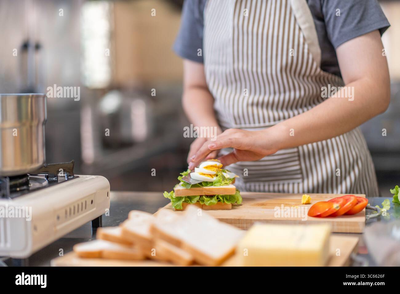 Jeune femme asiatique dans la vingtaine préparant un sandwich avec oeuf dur, laitue et tomate dans une cuisine commerciale. Idéal pour la préparation de la nourriture, la cuisine, culinaire Banque D'Images