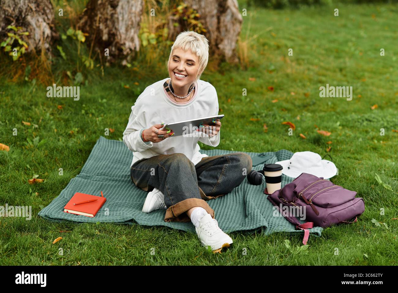 Charmante jeune femme se détend sur une couverture dans le parc, souriant et utilisant sa tablette avec joie. Banque D'Images
