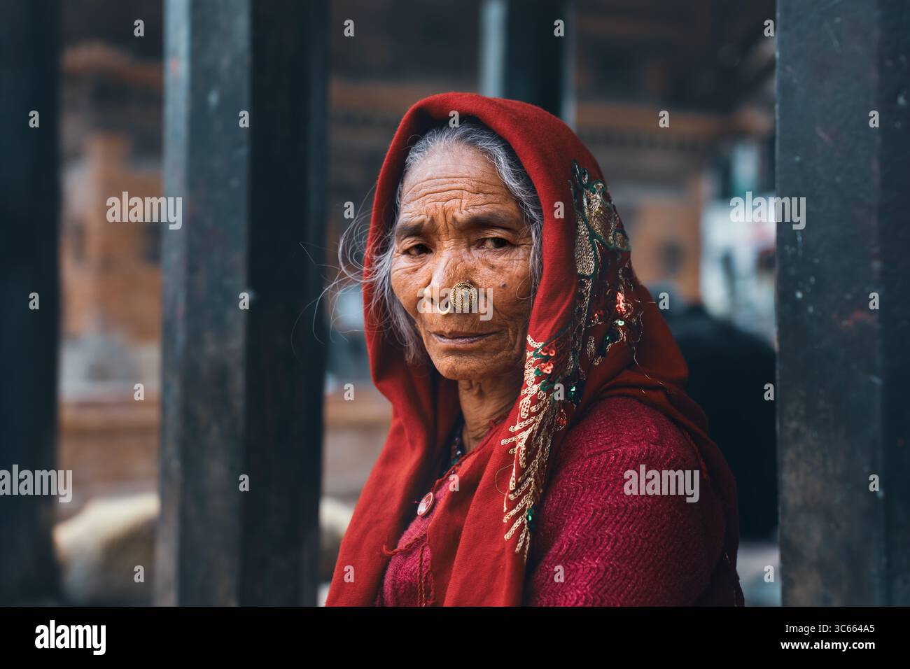 Dhangadhi, Népal - 03 mars 2023 : vue d'une femme âgée avec un châle rouge et un anneau nasal, debout devant une structure en bois altérée, son regard tenant des histoires de temps et de résilience. Banque D'Images
