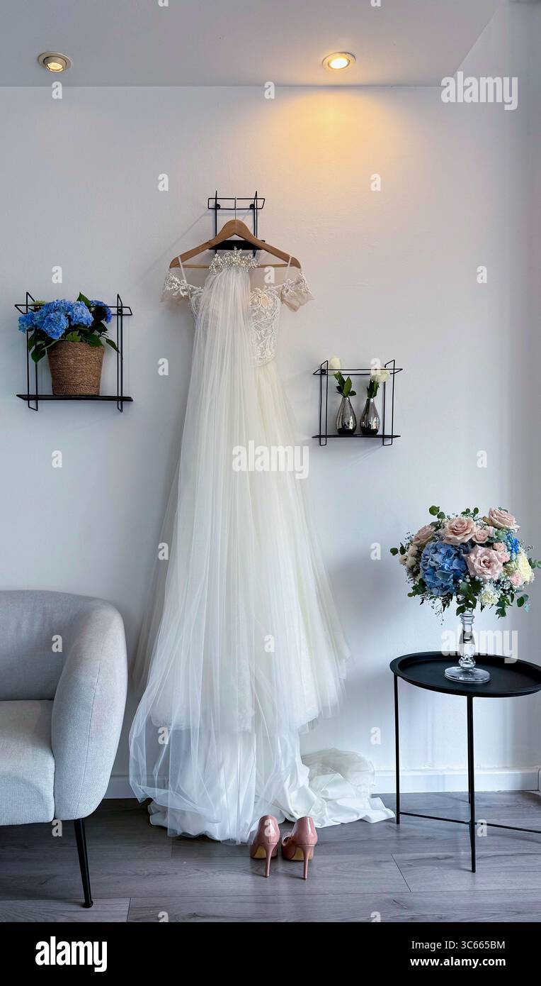 Élégante robe de mariée blanche accrochée à un cintre dans une pièce légèrement éclairée, préparée pour la célébration du mariage à venir Banque D'Images
