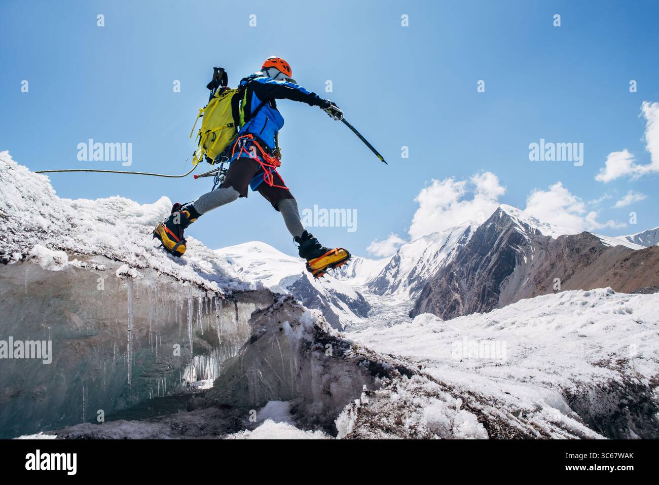Grimpeur en vitesse vibrante saute au-dessus de la crevasse sur le glacier glacé pendant l'ascension du pic de Lénine sous le ciel dégagé avec des sommets enneigés en arrière-plan. Acte extrême Banque D'Images