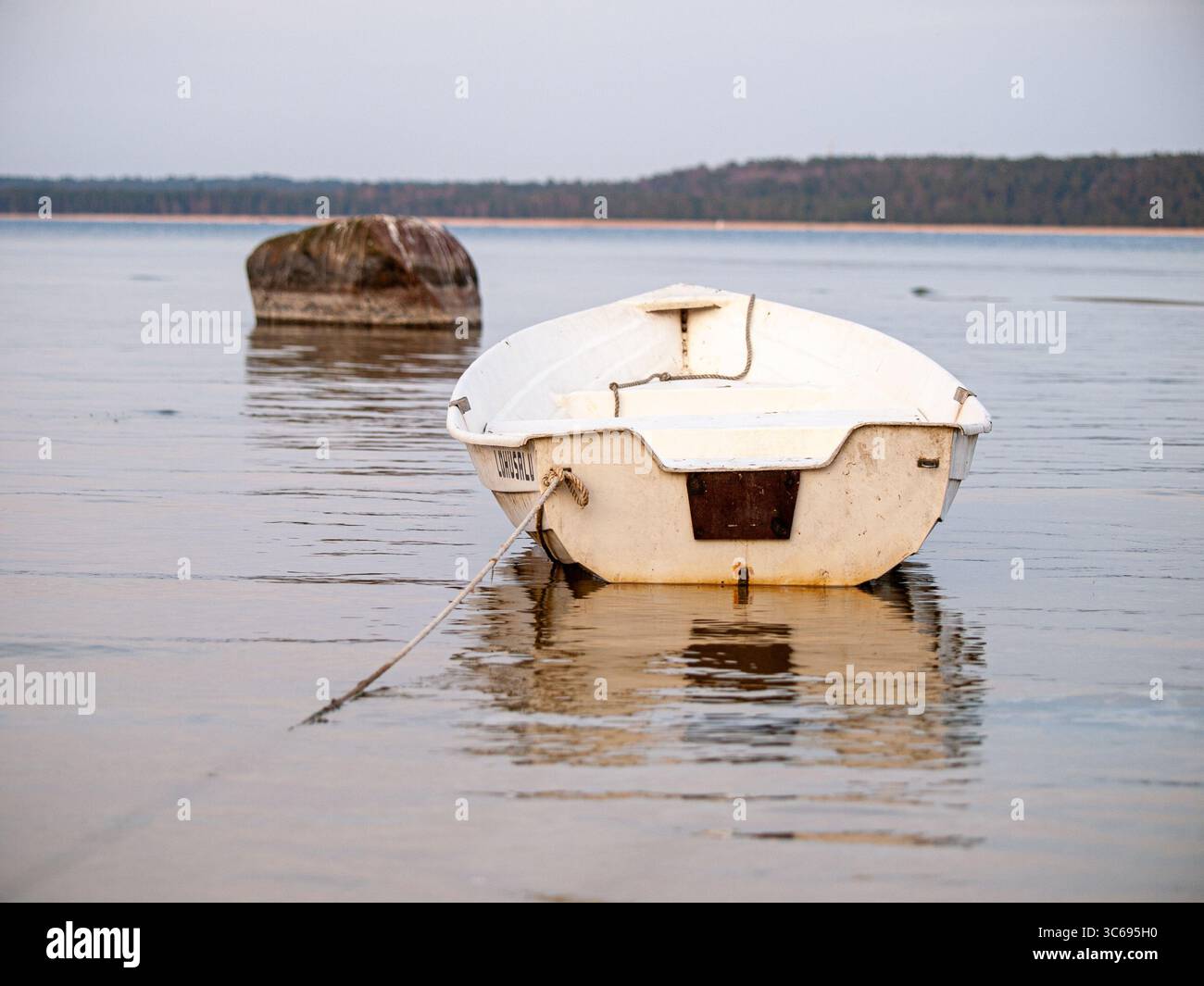 Un petit bateau en bois blanc ancré dans l'eau calme, créant des reflets sur la surface avec un gros rocher visible dans l'eau à proximité. Banque D'Images