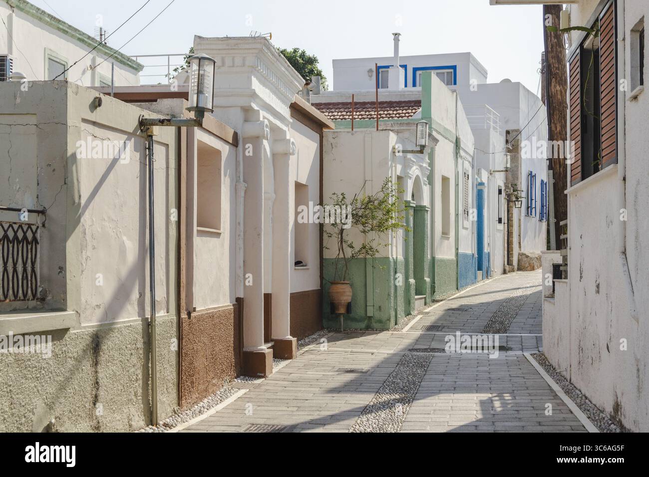 Vue sur la rue étroite baignée de soleil, bordée de bâtiments affichant des murs blancs, des éclats de couleur et des pavés à Koskinou, Rodhes, Grèce. Banque D'Images