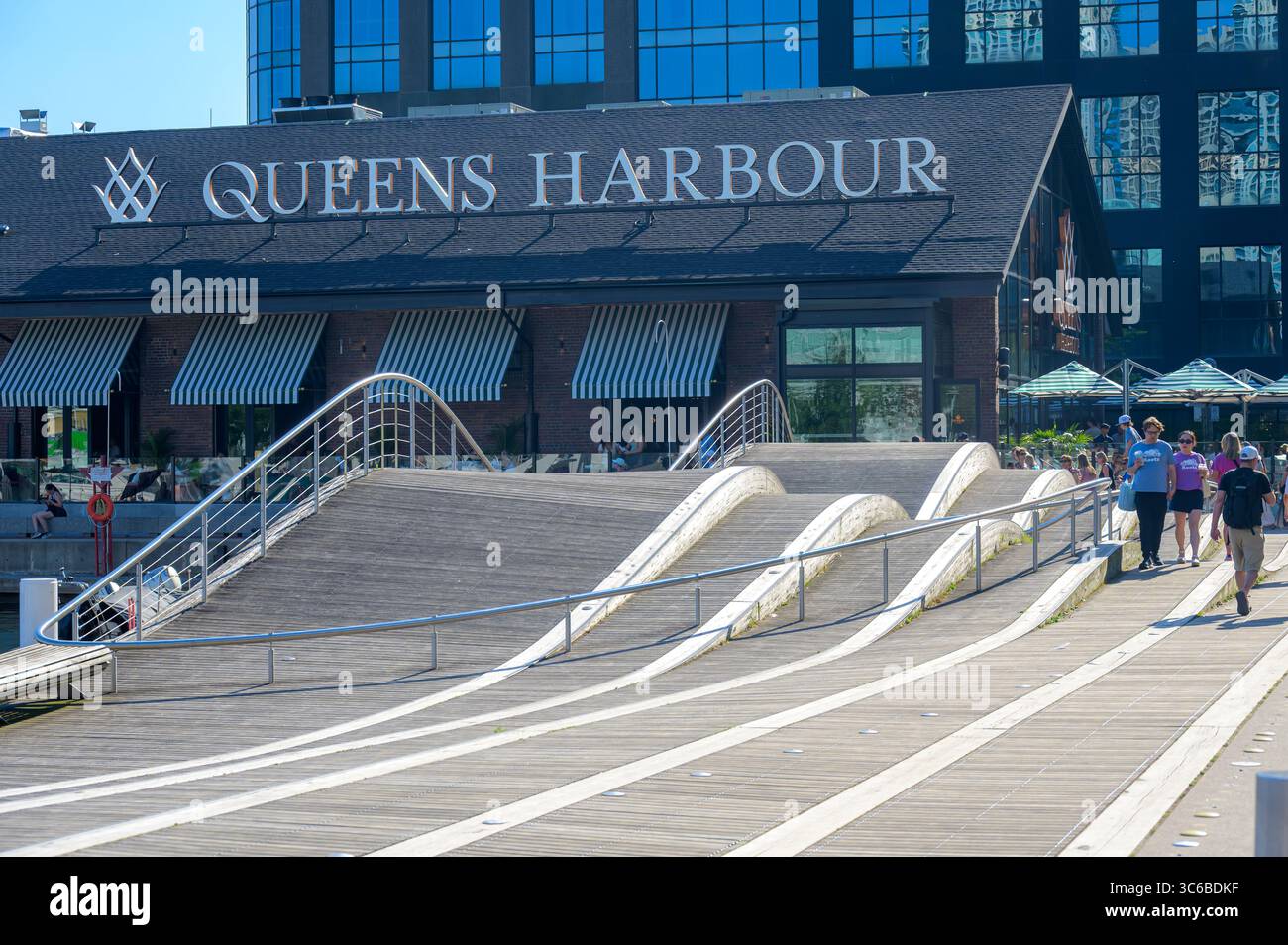 Les gens marchent à côté du bâtiment Queens Harbour près d'une promenade en forme de vague au bord de l'eau. Banque D'Images