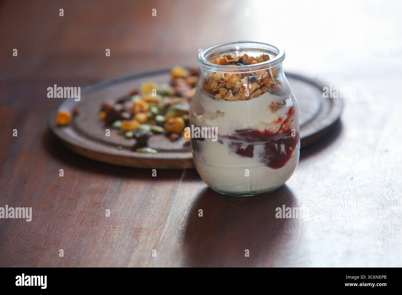 gros plan de pot en verre rempli de yaourt crémeux et d'un mélange en couches de granola croquant et de muesli, placé sur une table en bois avec des noix et des graines dispersées Banque D'Images