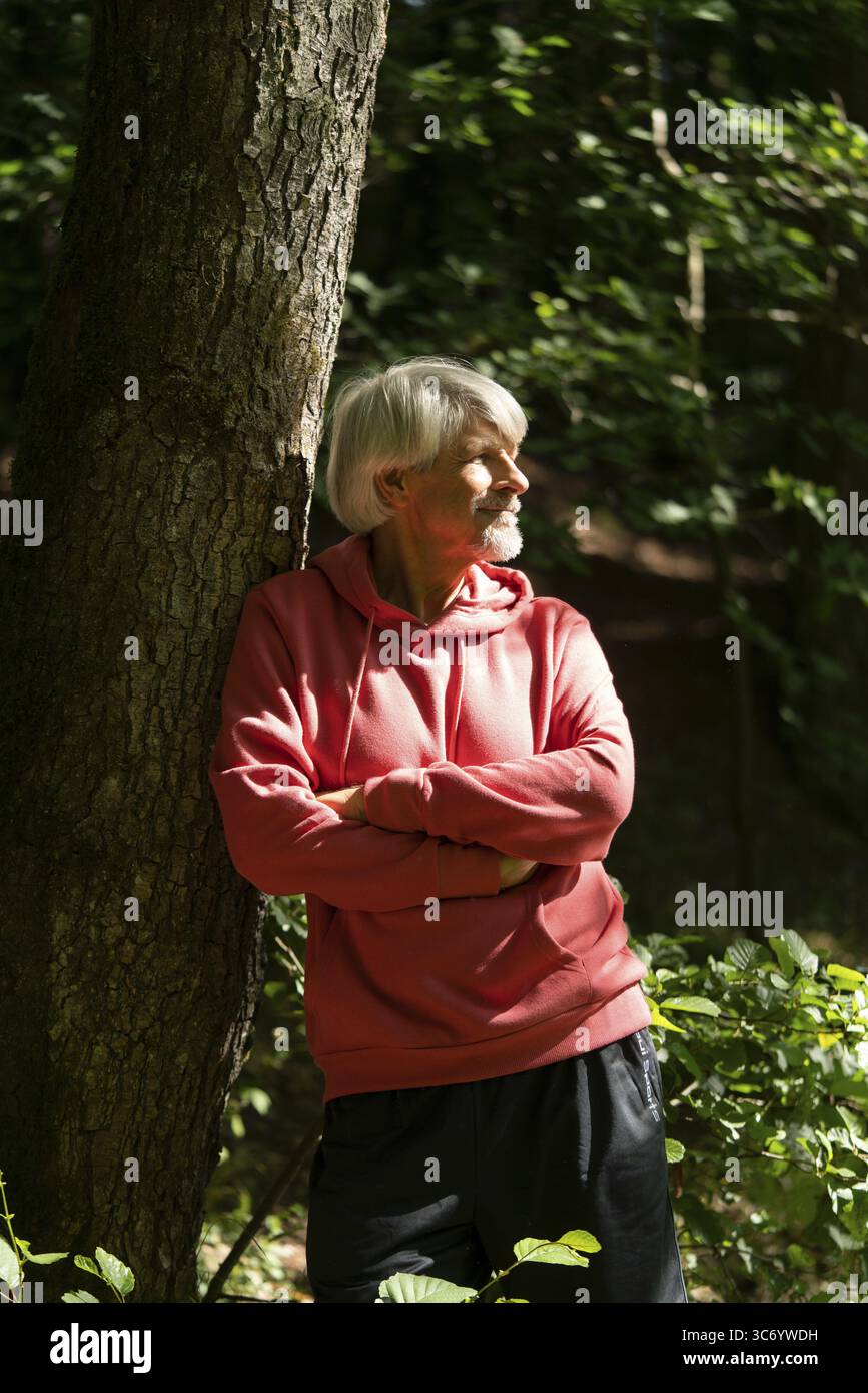 En forme, un vieil homme se penche contre un arbre dans la forêt et regarde au loin, Trimm dich Pfad Banque D'Images