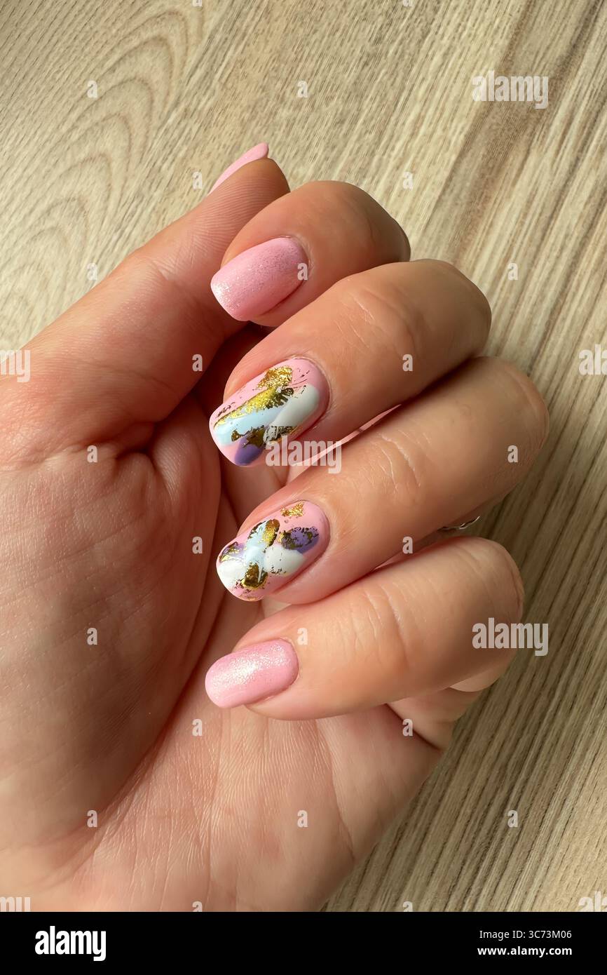 gros plan d'une main féminine avec une manucure rose chic et deux clous d'accent avec un design abstrait unique avec des feuilles blanches, violettes et dorées Banque D'Images